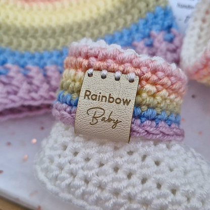 A close-up of a handmade crocheted baby bootie with pastel rainbow stripes in pink, yellow, blue, and purple, featuring a beige faux-leather strap with a 'Rainbow Baby' faux leather tag.