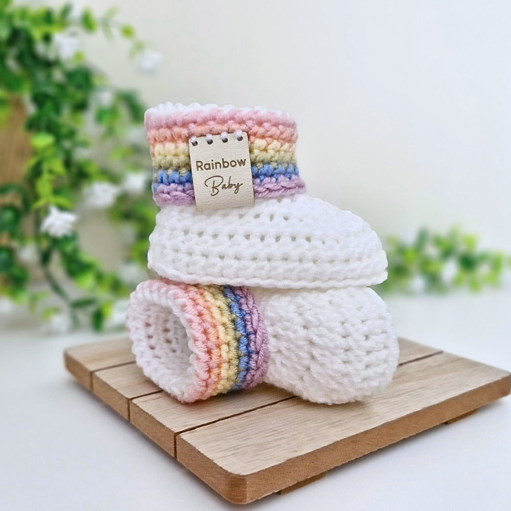 A pair of handmade crocheted baby booties stacked on a small wooden block. The booties are primarily white with a pastel rainbow stripe pattern around the cuffs, finished with a 'Rainbow Baby' faux leather tag 