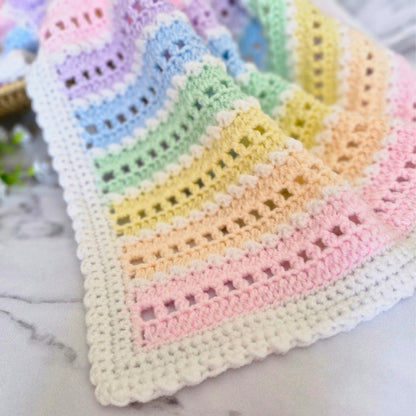 A pastel rainbow crocheted baby blanket with a white scalloped border, laid out on a light marble surface.