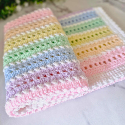 A handmade crocheted baby blanket with pastel rainbow stripes in pink, yellow, green, blue, and purple, folded on a light marble surface.