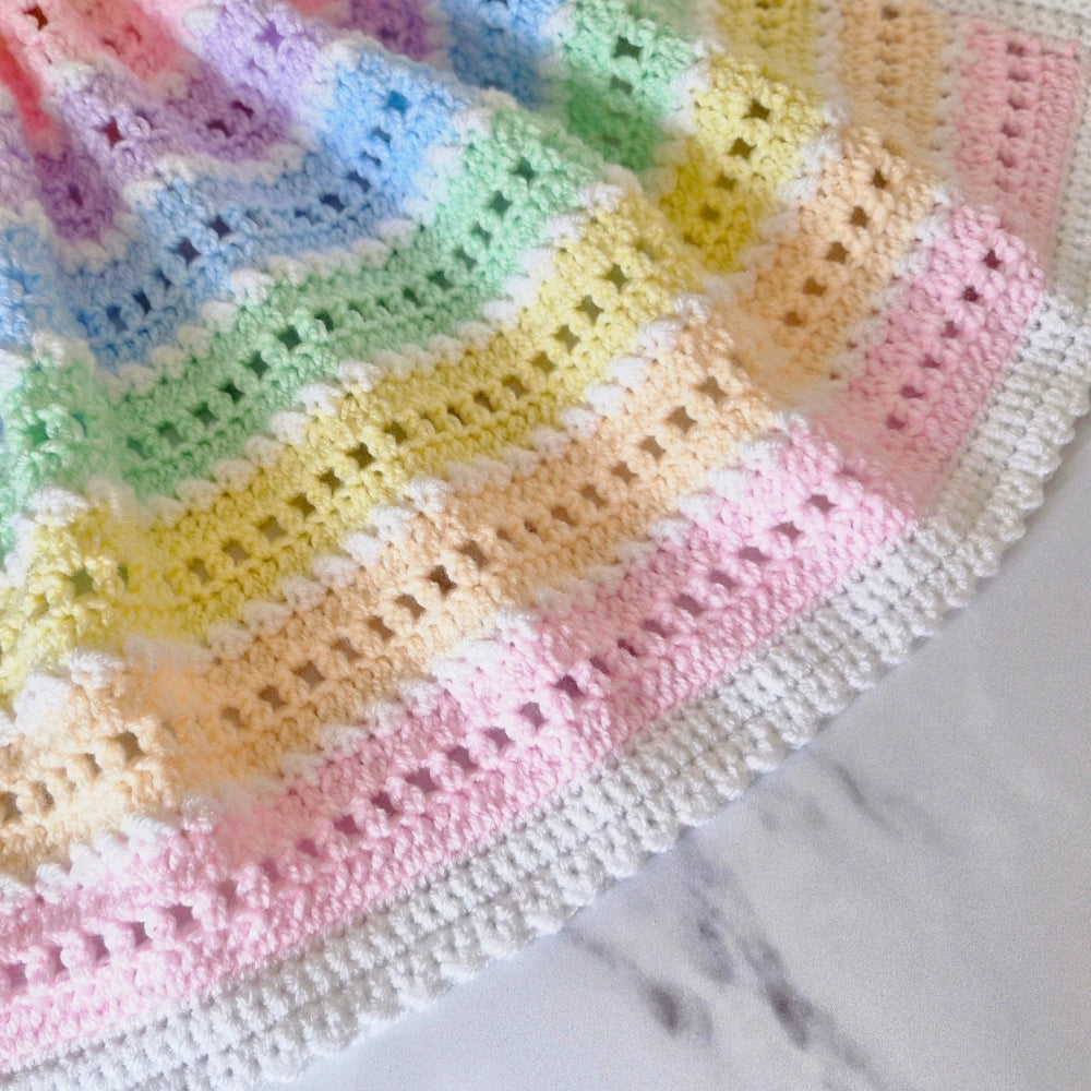 A close-up of a crocheted blanket with pastel rainbow stripes in pink, blue, green, yellow, and peach, finished with a white scalloped border, laid on a light marble surface.