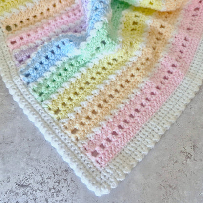 A close-up of a crocheted baby blanket with pastel rainbow stripes and a cream border, laid on a textured gray surface.