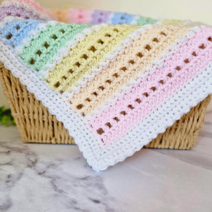 A pastel crocheted baby blanket with rainbow-colored stripes draped over a woven wicker basket on a marble surface.