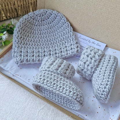 Grey crocheted baby hat, booties, and mittens set on a white surface with a card in the background.