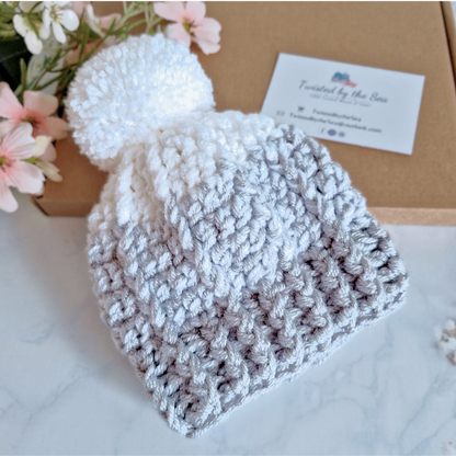 Crochet baby hat with a pom-pom on a white surface with flowers and a business card in the background.