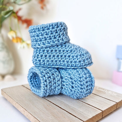 Pair of blue crochet baby booties on a wooden surface with a blurred floral background.