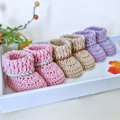 Set of summerween inspired crocheted baby booties in pink, beige, and purple on a white surface with a blurred background.