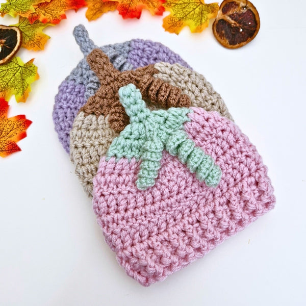 Three crocheted summerween beanies in different colors with a green leaf decoration on a white background with autumn leaves.
