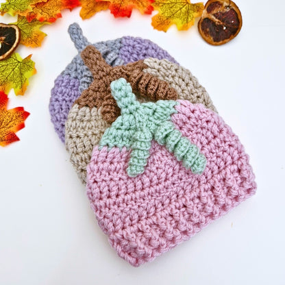 Three crocheted summerween beanies in different colors with a green leaf decoration on a white background with autumn leaves.