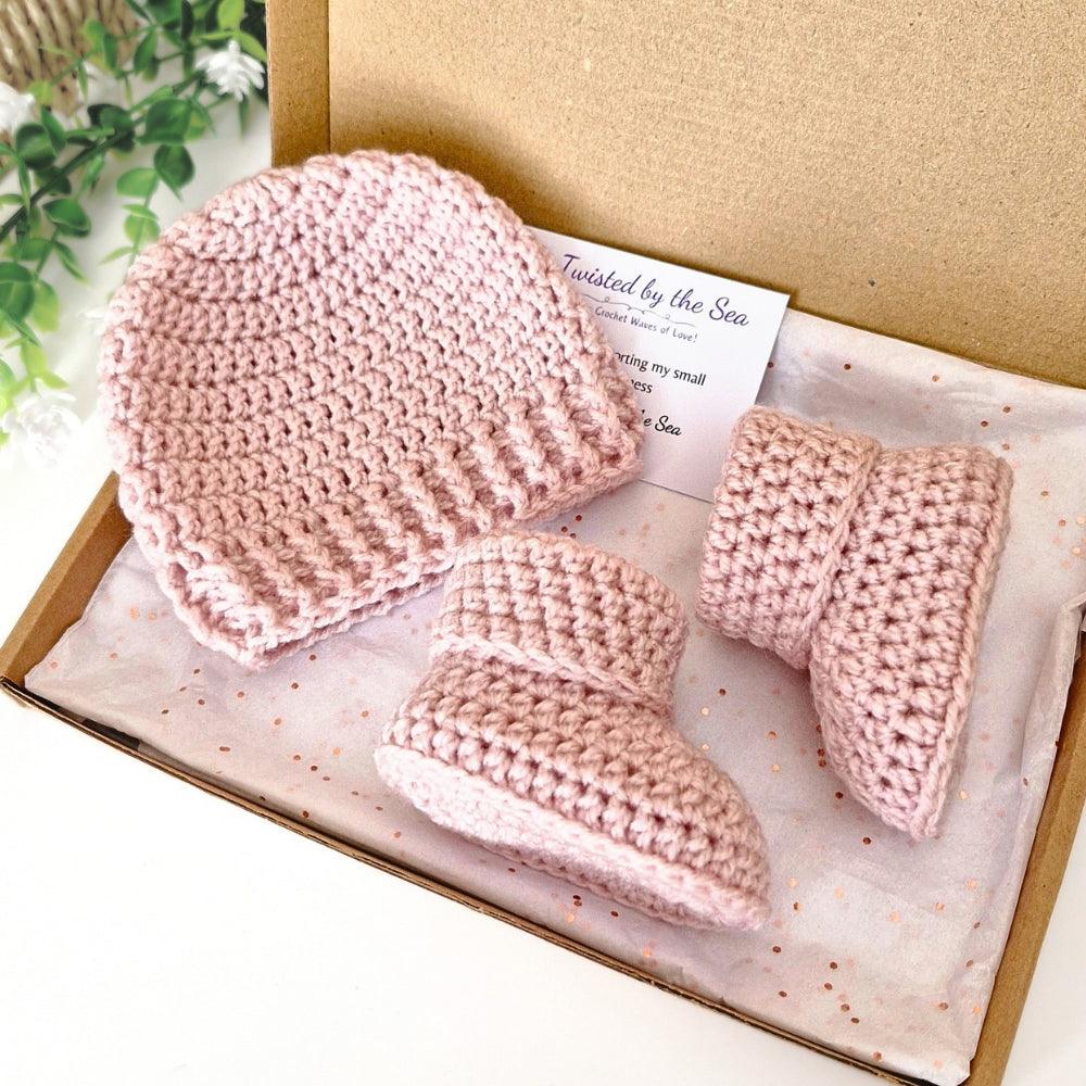 Pink crocheted baby hat and booties set in a box with a card on a white background