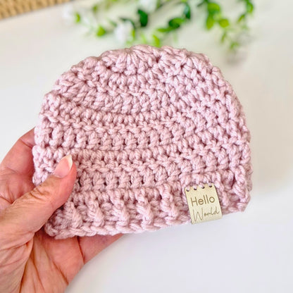 Pink crocheted baby hat with 'Hello World' tag held by a hand on a light background