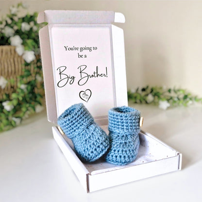 Pair of blue baby booties in a box with a card that reads 'You're going to be a Big Brother!' on a white surface with floral background.