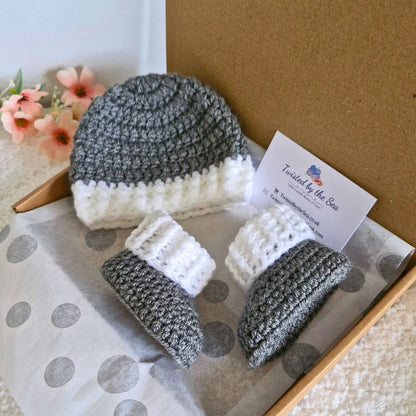A handmade grey and white crochet baby hat and matching booties, displayed in a letterbox-friendly gift box with tissue paper.