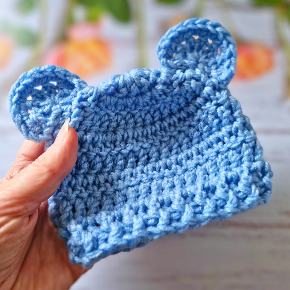 Blue crocheted bear hat held by a hand with a blurred background