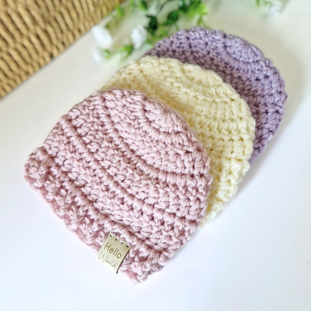 Three crochet hats in pink, cream, and purple on a white surface with a floral background.