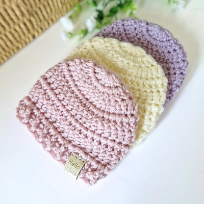 Three crochet hats in pink, cream, and purple on a white surface with a floral background.