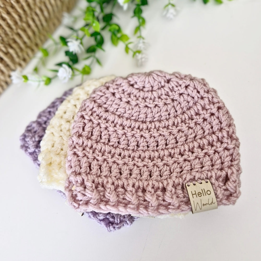 Three crocheted baby hats, one pink and one cream and one purple, on a white surface with greenery in the background.