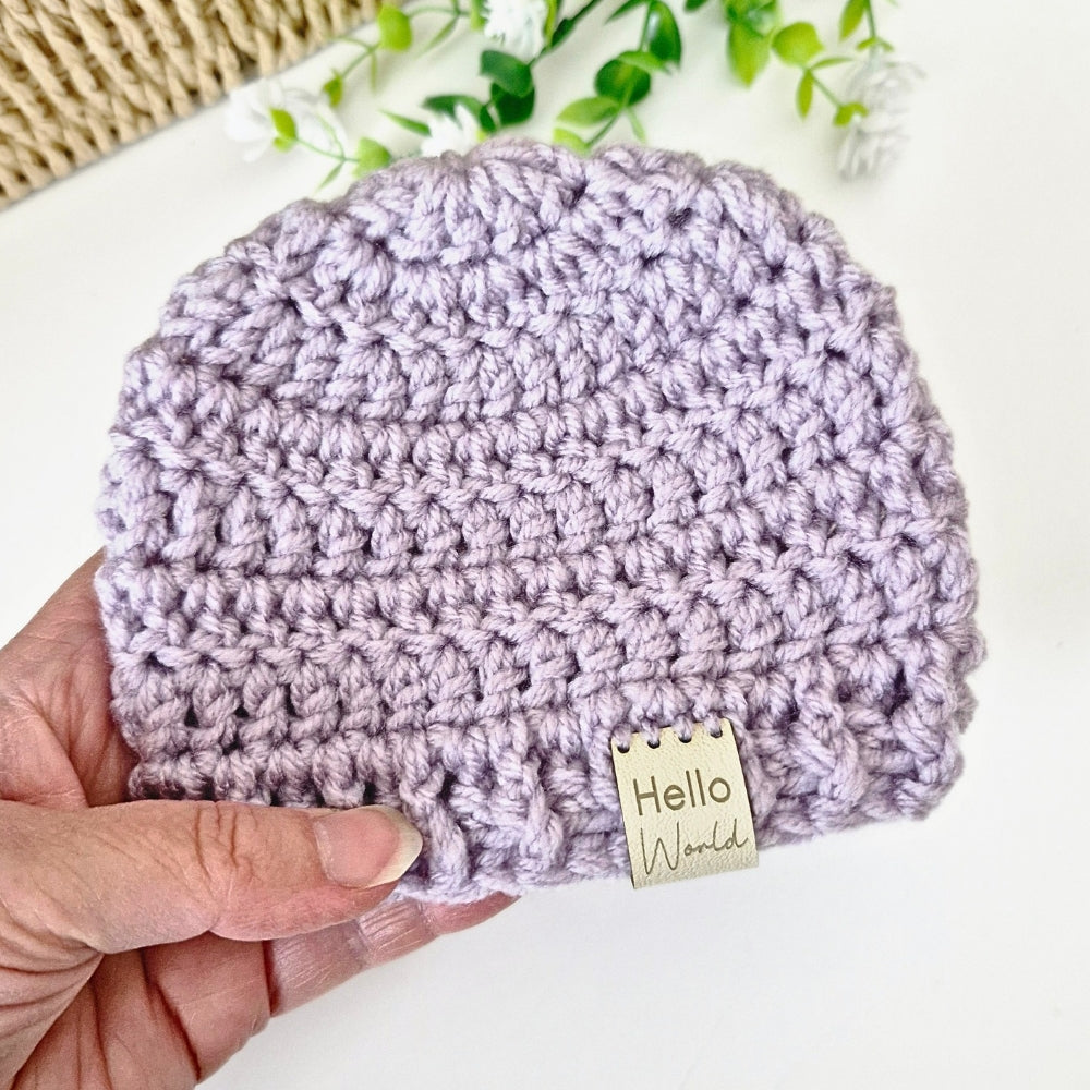 Hand holding a purple crocheted beanie with 'Hello World' tag on a white background