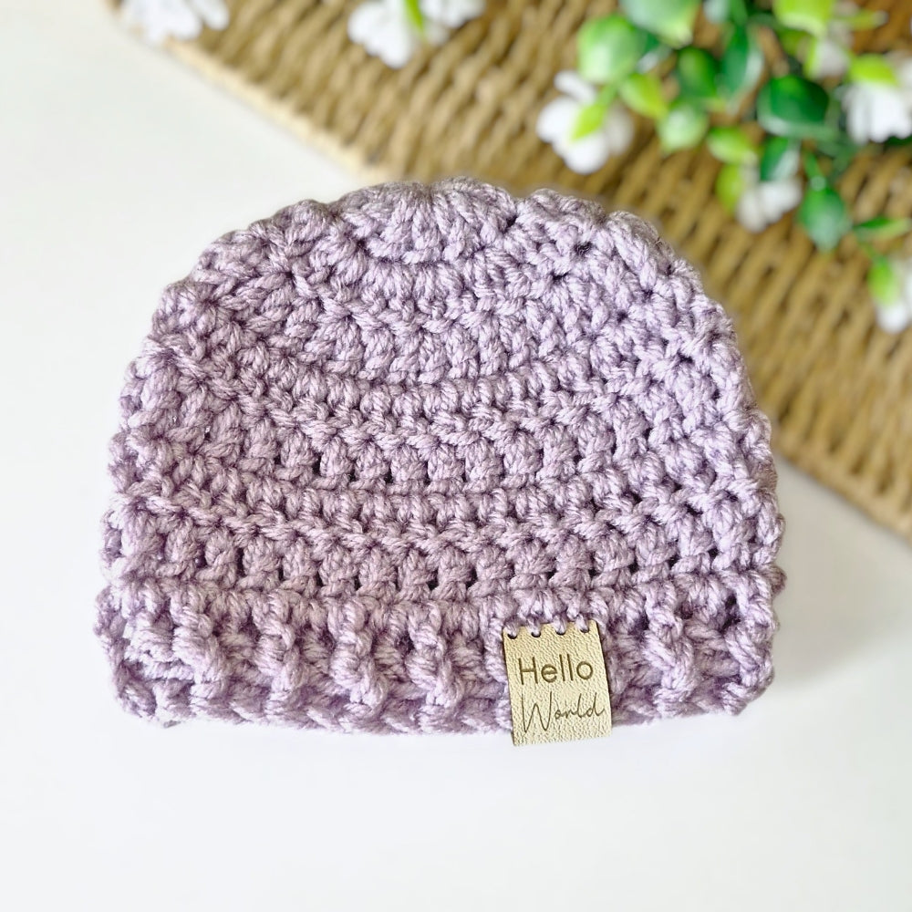 Crocheted purple beanie with a 'Hello World' tag on a white surface.