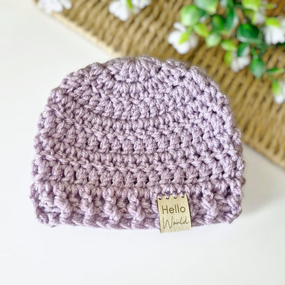 Crocheted purple beanie with a 'Hello World' tag on a white surface.