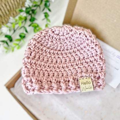 Pink crocheted baby hat with 'Hello World' tag on a light background