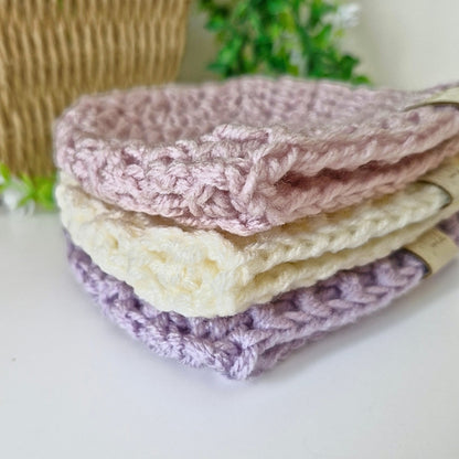 Stack of crocheted baby hats in pink, cream, and purple on a white surface with a blurred background.