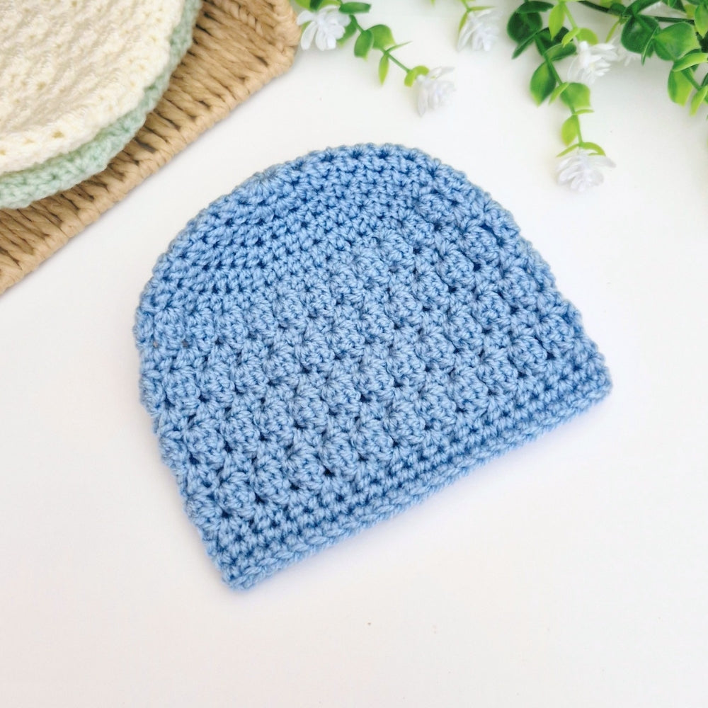Blue crocheted beanie on a white surface with a basket and greenery in the background