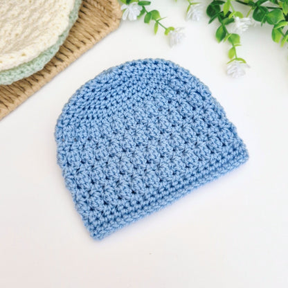Blue crocheted beanie on a white surface with a basket and greenery in the background