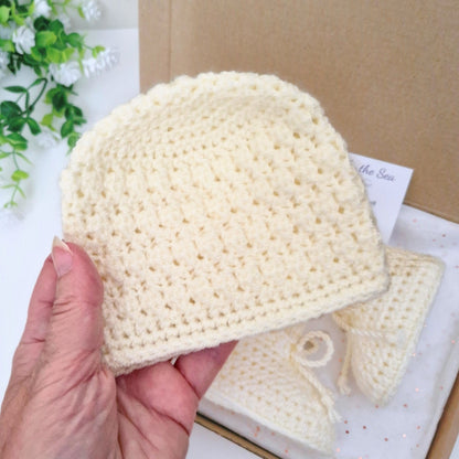 Hand holding a cream crochet baby hat with matching booties on a light background