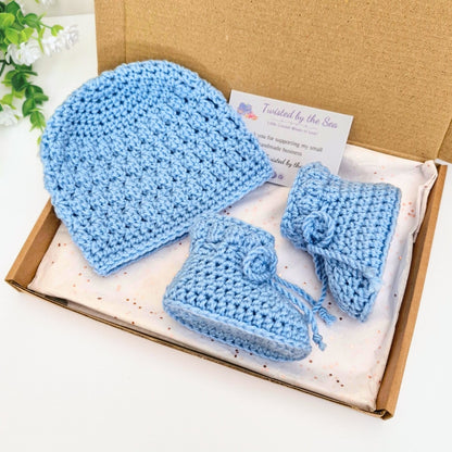 Set of blue crocheted matching baby hat and a booties in a cardboard box with a plant in the background.