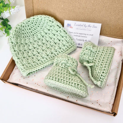 Set of tea green crocheted baby matching hat and booties in a box with a card on a white background