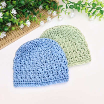 Two crocheted hats, one blue and one green, on a white surface with greenery in the background.