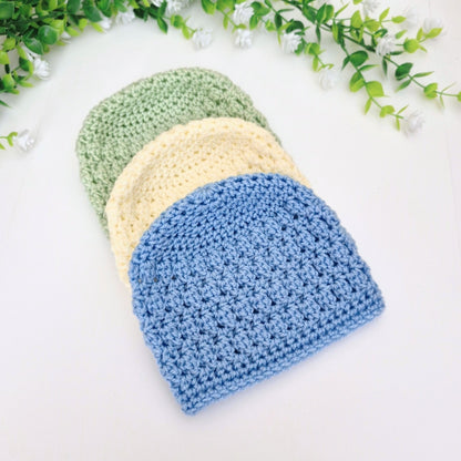 Three crocheted beanies in green, cream, and blue on a white background with greenery.