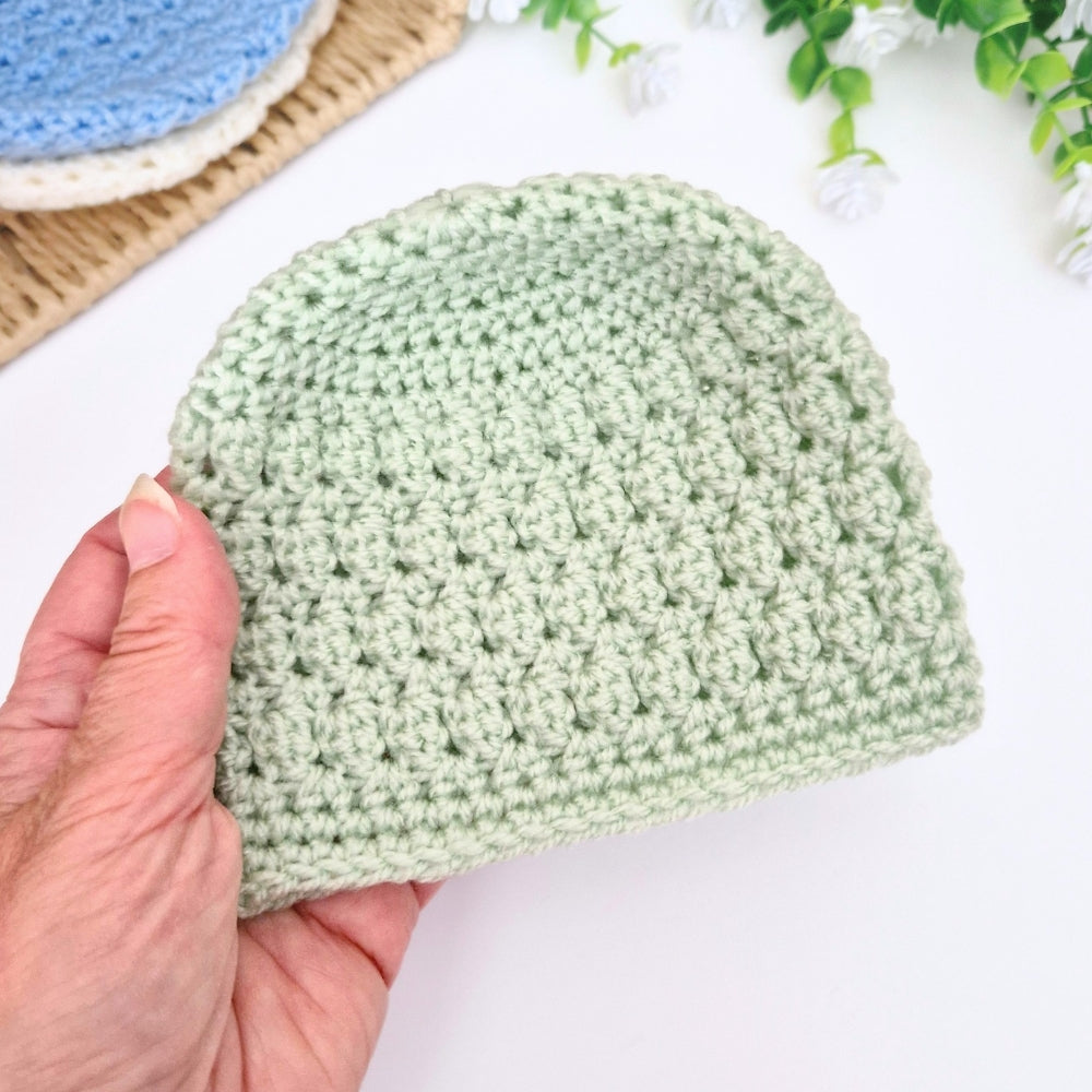 Hand holding a green crocheted beanie on a white surface with a blurred background
