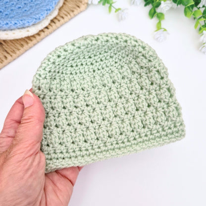 Hand holding a green crocheted beanie on a white surface with a blurred background