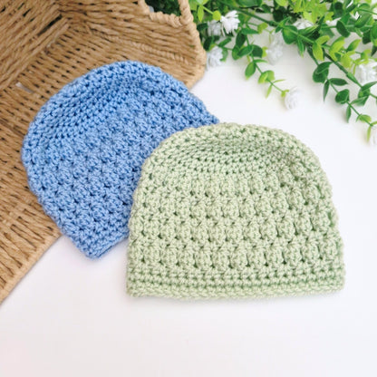 Two crocheted hats, one blue and one green, on a white surface with a basket and greenery in the background