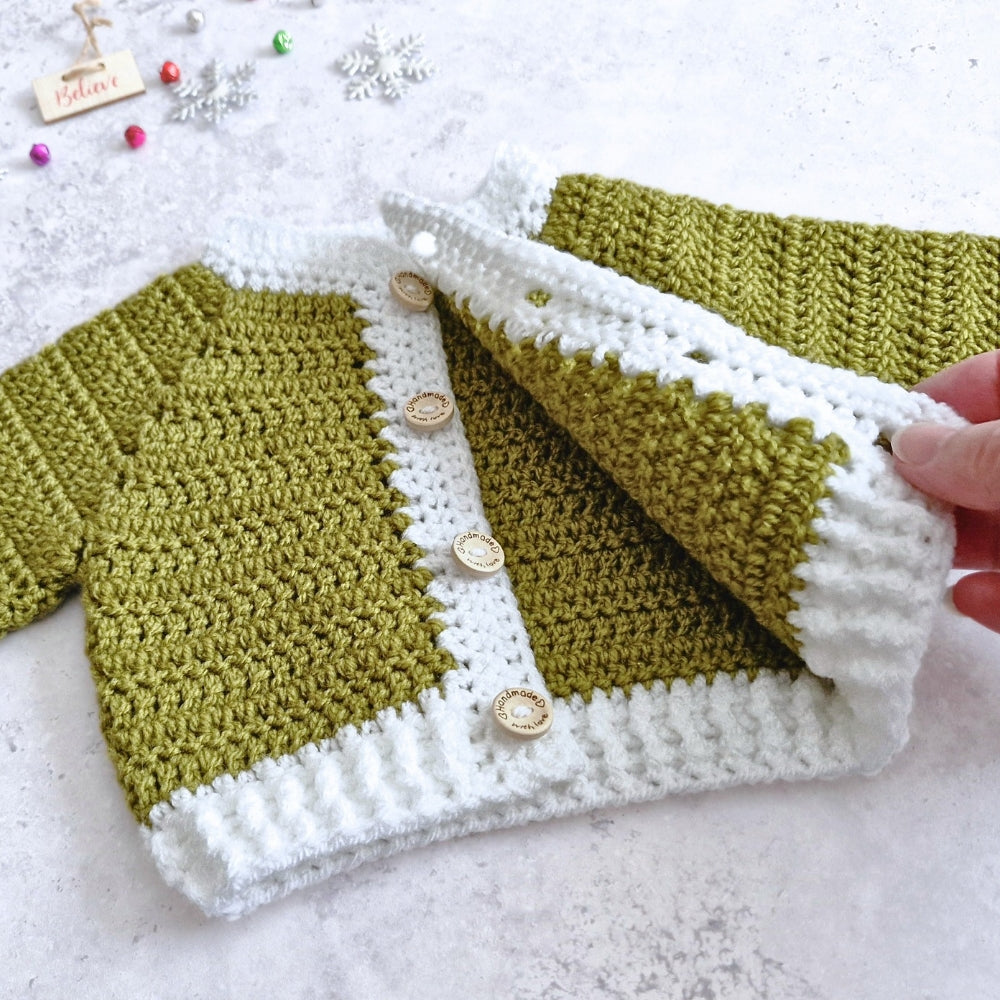 A handmade crocheted cardigan in mistletoe green and white, featuring decorative wooden buttons down the front. A hand is holding up the right side of the cardigan to show the texture and construction.