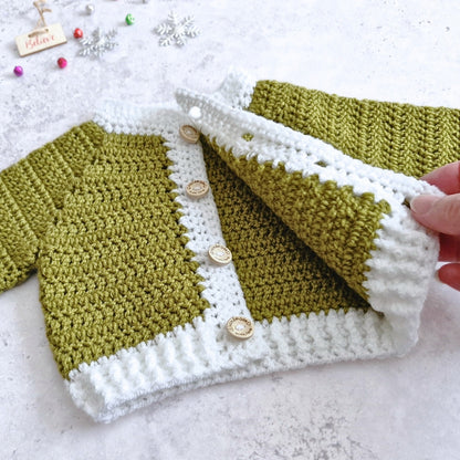 A handmade crocheted cardigan in mistletoe green and white, featuring decorative wooden buttons down the front. A hand is holding up the right side of the cardigan to show the texture and construction.