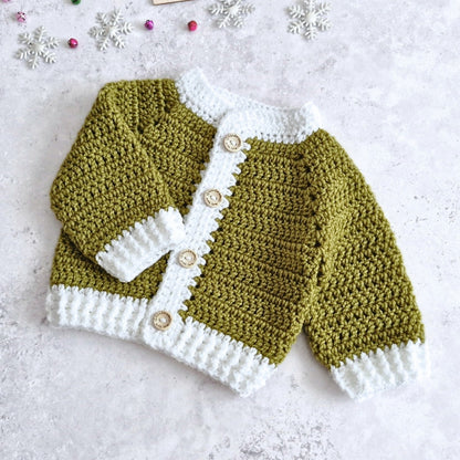 A handmade crocheted baby cardigan sweater in mistletoe green and white, featuring three wooden buttons, laid flat on a textured light gray surface with small decorative snowflakes.
