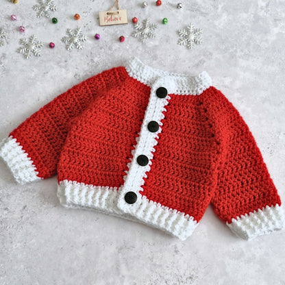 A red and white crocheted baby cardigan styled like a Santa hat, featuring black buttons down the front, laid flat on a light gray textured surface with holiday decorations.